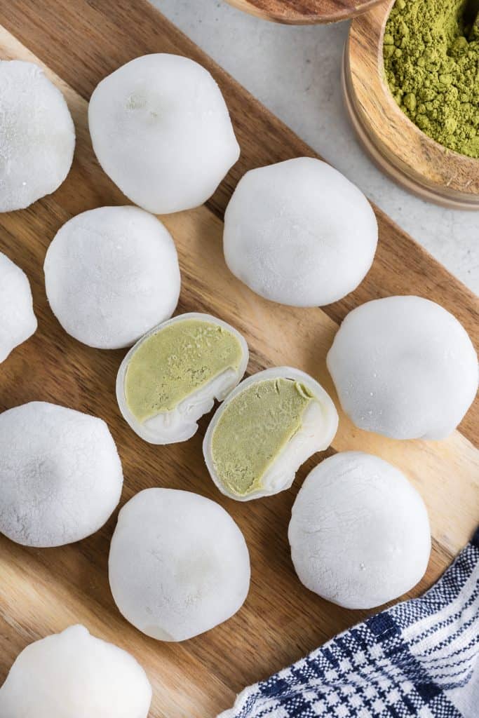 Overhead shot of a wooden cutting board covered in rows of white mochi, with one in the middle which has been cut in two revealing the green tea ice cream inside. The upper right corner has a wooden jar of green tea powder peeking in.