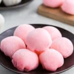 A close up head on shot of a small black plate with 7 pink ice cream mochis piled in a circle ready to serve. In the background on the left is a small plate filled with white ice cream mochis and on the right is a wood block with two pink ice cream mochis.