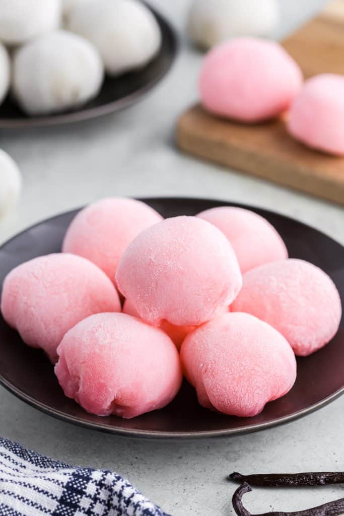 A close up head on shot of a small black plate with 7 pink ice cream mochis piled in a circle ready to serve.  In the background on the left is a small plate filled with white ice cream mochis and on the right is a wood block with two pink ice cream mochis.