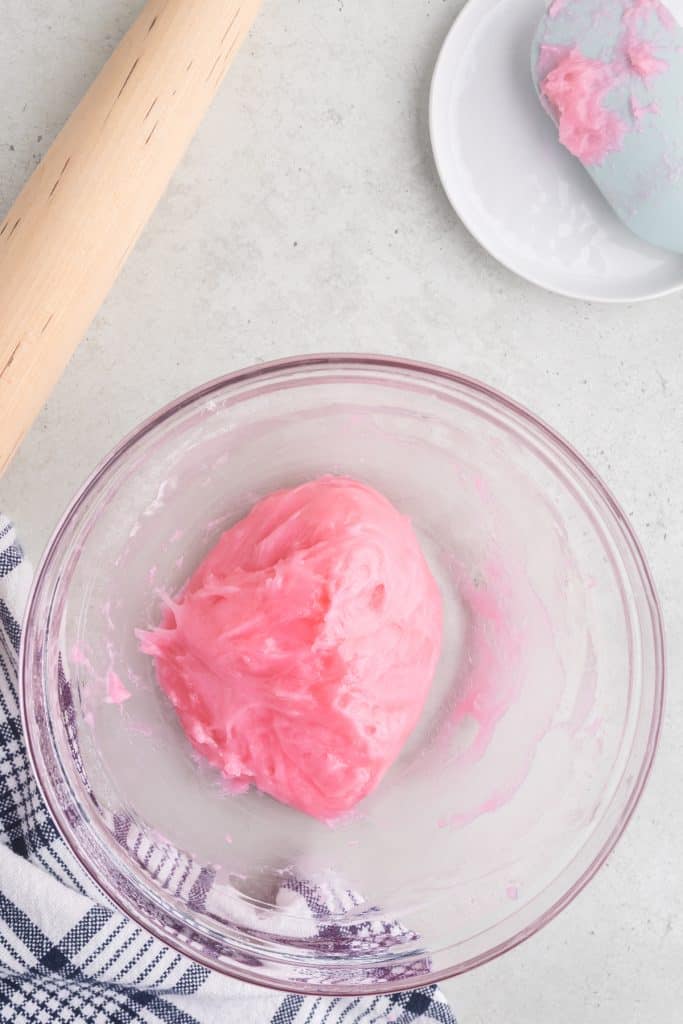 overhead shot of a mixing bowl filled with the cooked pink mochi dough. The upper left corner has a rolling pin and the upper right corner has blue rubber spatula spoon with bits of pink mochi dough stuck on i it.