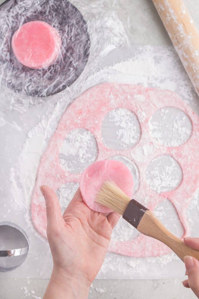 overhead picture of pink mochi dough which has been rolled out and circles cut out. One circle of dough is being held in a hand while the other hand dusts off excess cornstarch with a pastry brush.