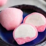 45 degree angle shot close up on a small blue plate with one whole pink ice cream mochi and and another pink mochi which has been cut in half and opened up to reveal white vanilla ice cream inside.