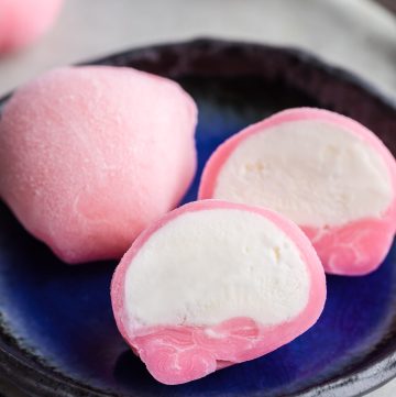 45 degree angle shot close up on a small blue plate with one whole pink ice cream mochi and and another pink mochi which has been cut in half and opened up to reveal white vanilla ice cream inside.