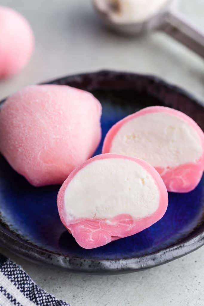 45 degree angle shot close up on a small blue plate with one whole pink ice cream mochi and and another pink mochi which has been cut in half and opened up to reveal white vanilla ice cream inside.