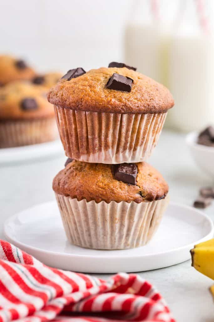 side angle shot of two banana chocolate chunk muffins stacked on top of each other. In the background is a plate of more muffins and two glasses of milk with red striped straws. In the foreground you can see some chocolate and bananas peeking in to the frame