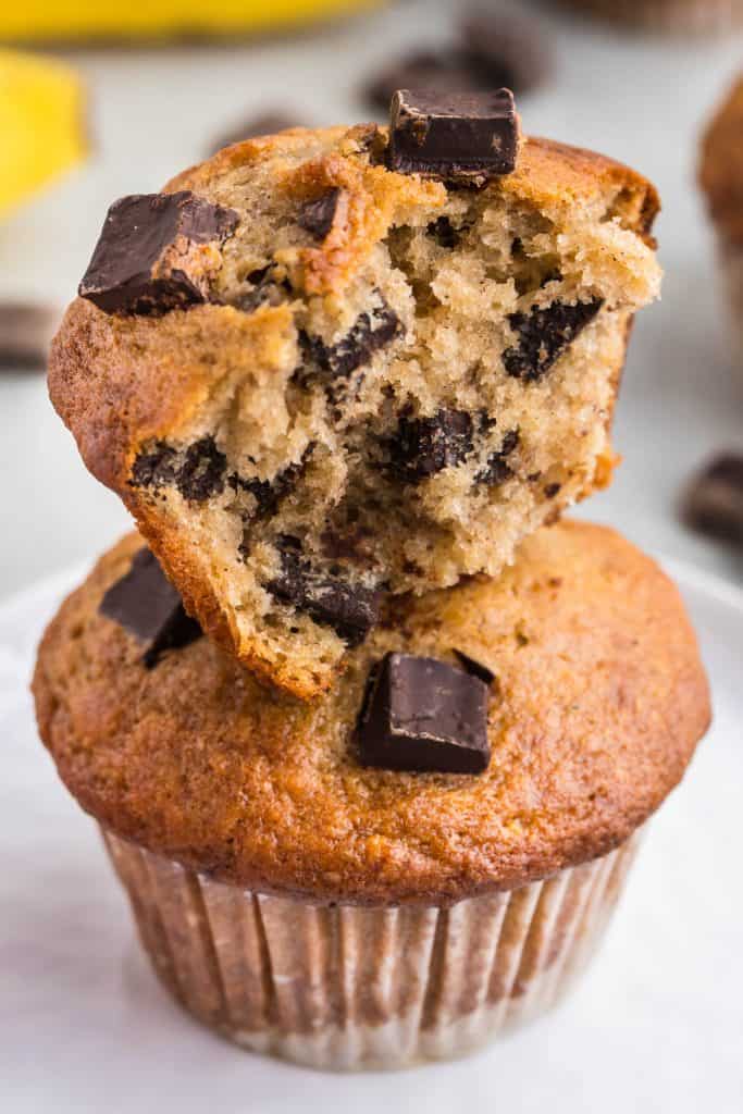 Close up view of a torn open half banana chocolate chunk muffin stacked on top of another muffin. Lots of chocolate chunks can be seen in the interior.