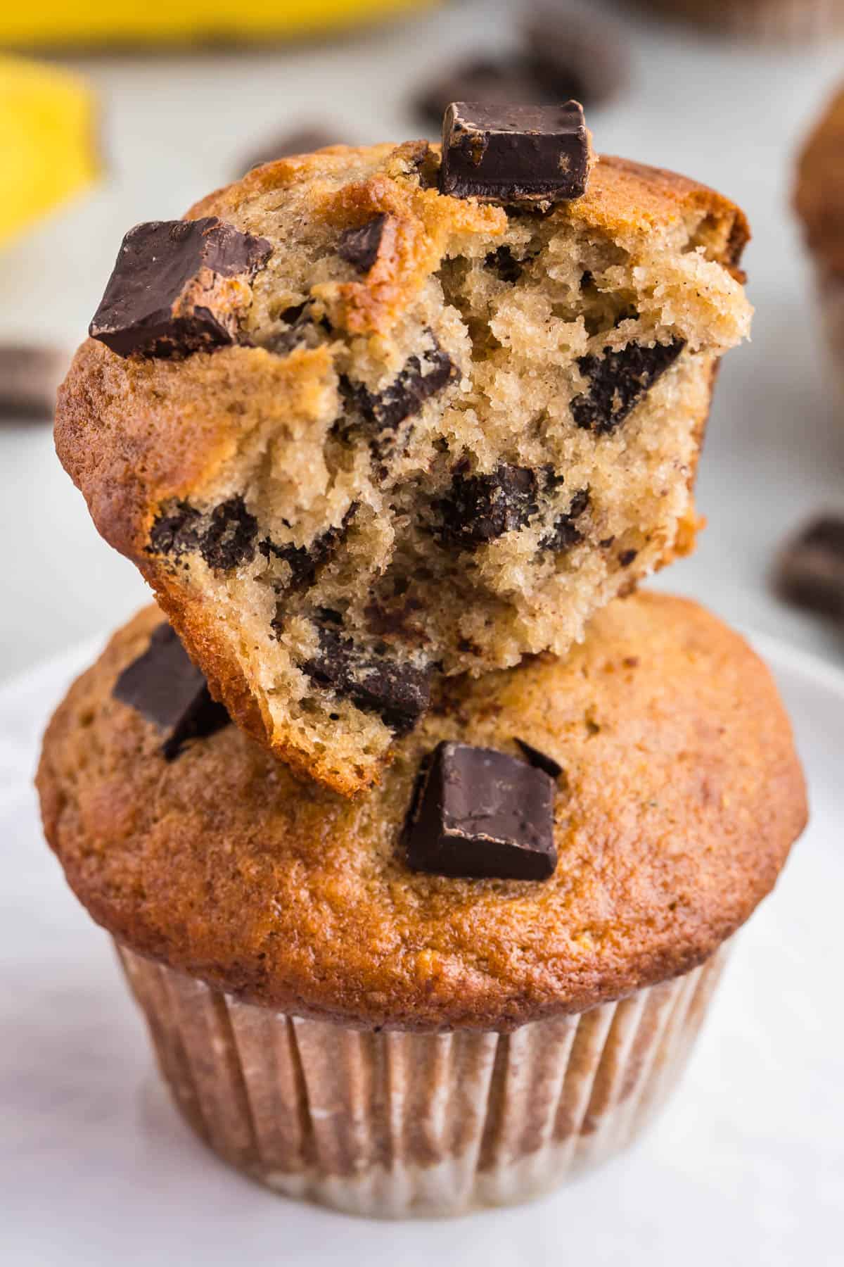 Close up view of a torn open half banana chocolate chunk muffin stacked on top of another muffin. Lots of chocolate chunks can be seen in the interior.