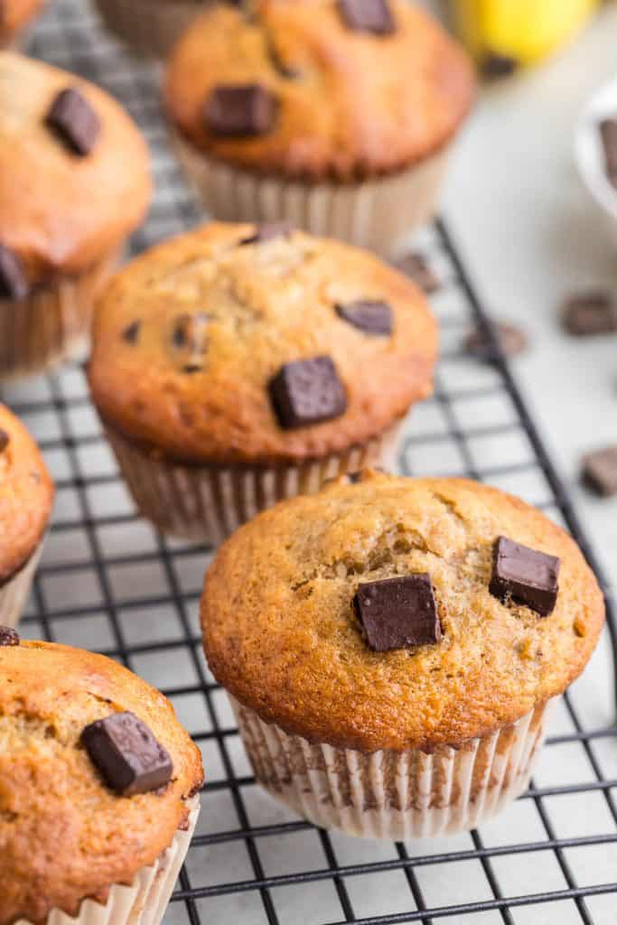 45 degree angle shot of banana chocolate chunk muffins cooling on a cooling rack. Chocolate chunks are scattered along the side of the cooling rack as well as a couple of bananas peeking in at the top right of the picture.