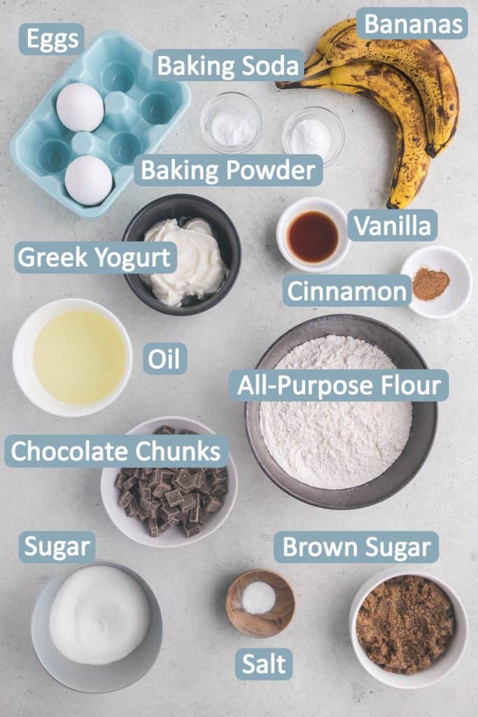 Overhead shot of all the ingredients, labelled, necessary to make Banana Chocolate Chunk Muffins. Ingredients are from top to bottom: eggs, baking soda, baking powder, bananas, Greek yogurt, vanilla, cinnamon, oil, all-purpose flour, chocolate chunks, sugar, salt, and brown sugar