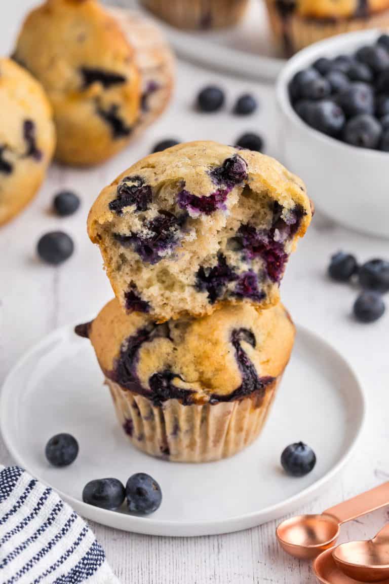 side shot of two cottage cheese blueberry muffins on a small white plate. One muffin has been pulled apart and is sitting atop the other muffin on the plate. You can see lots of blueberries inside the muffin. In the background there are more muffins and fresh blueberries scattered about.