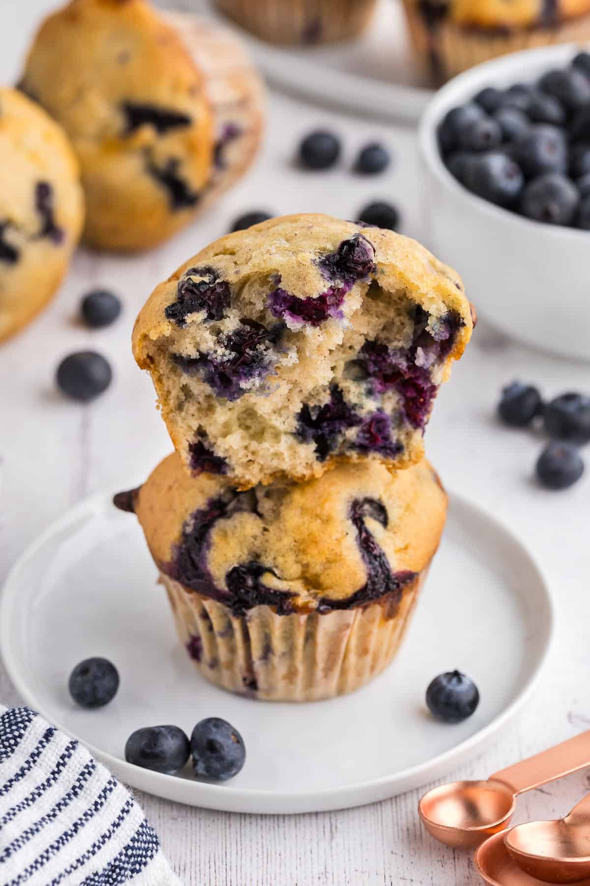 side shot of two cottage cheese blueberry muffins on a small white plate. One muffin has been pulled apart and is sitting atop the other muffin on the plate. You can see lots of blueberries inside the muffin. In the background there are more muffins and fresh blueberries scattered about.