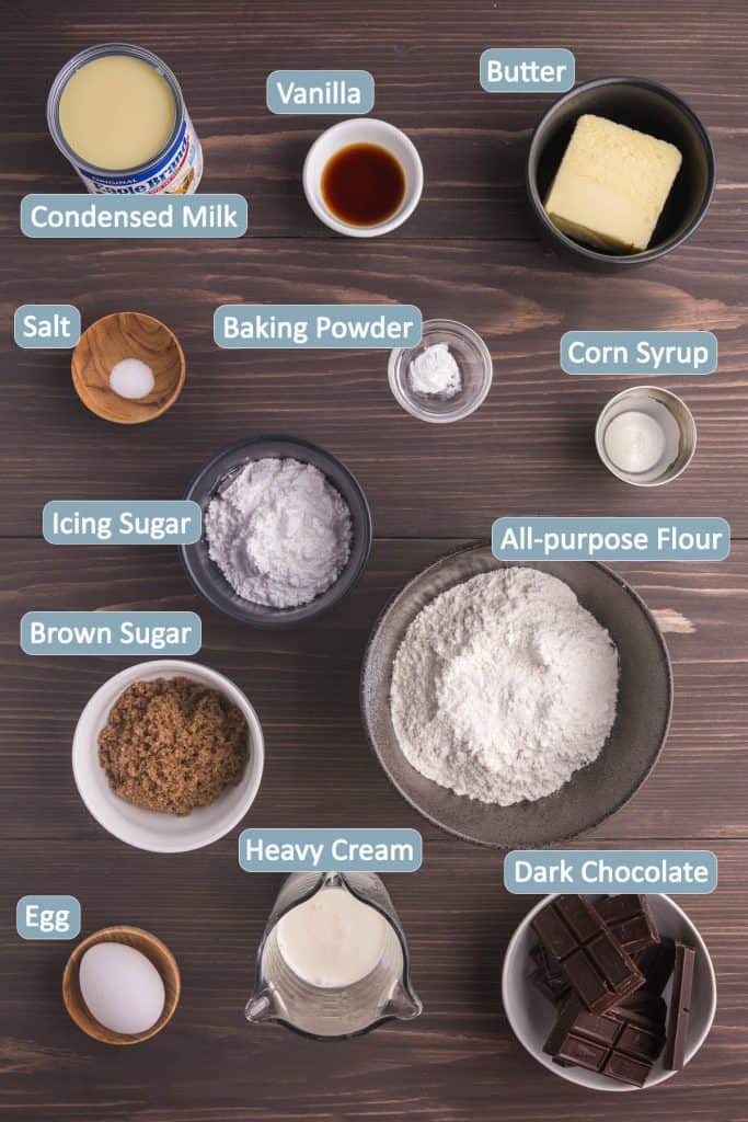 Overhead shot of ingredients needed to make a chocolate caramel tart. Ingredients are: condensed milk, vanilla extract, butter, salt, baking powder, corn syrup, icing sugar, all-purpose flour, brown sugar, egg, heavy cream, and dark chocolate