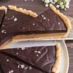 low 45 degree angle picture of a slice of chocolate caramel tart being lifted up and away from the whole tart. A small bottle of milk is peeking in in the upper left corner, and a small bunch of baby's breath flowers are in the upper right corner.