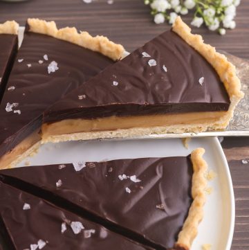 low 45 degree angle picture of a slice of chocolate caramel tart being lifted up and away from the whole tart. A small bottle of milk is peeking in in the upper left corner, and a small bunch of baby's breath flowers are in the upper right corner.