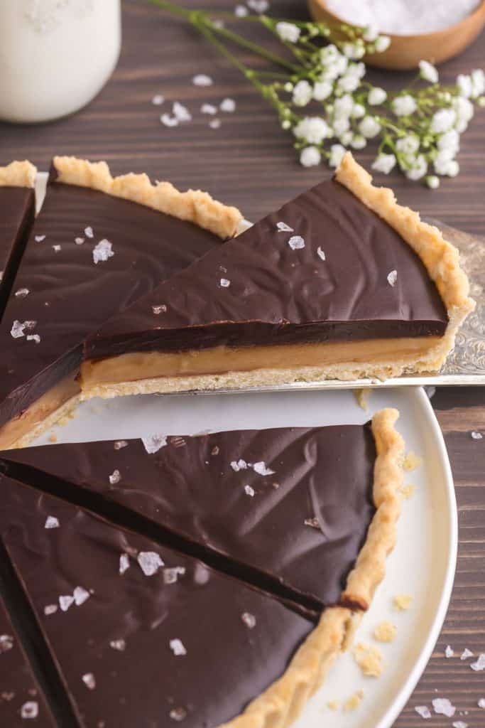 low 45 degree angle picture of a slice of chocolate caramel tart being lifted up and away from the whole tart. A small bottle of milk is peeking in in the upper left corner, and a small bunch of baby's breath flowers are in the upper right corner.