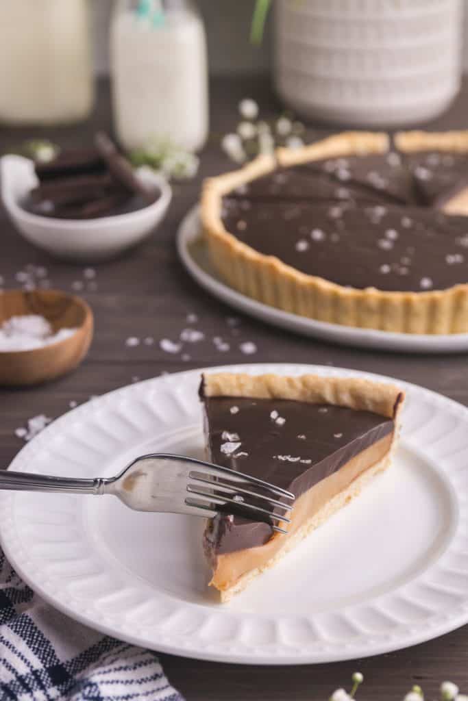 45 degree shot of a slice of chocolate caramel tart facing you, with a fork cutting in to the tip of the slice. In the background on the right is the rest of the tart, and on the left in the background is a small bowl of flaked sea salt and another small bowl of chocolate chunks