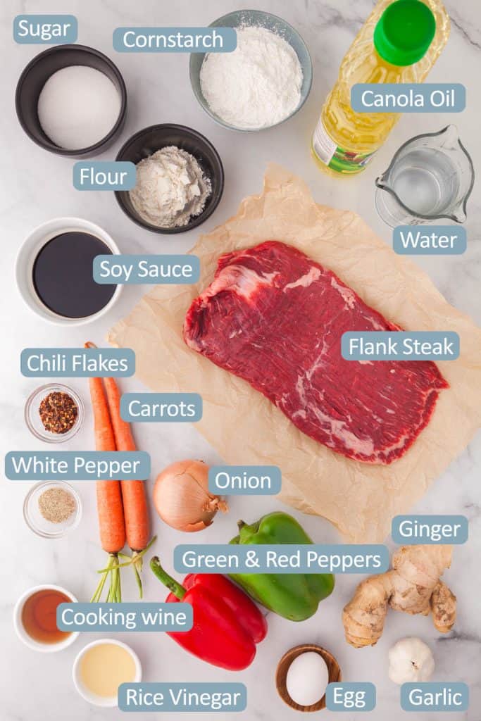 Overhead shot of all the ingredients used in making ginger beef. Ingredients include: flour, sugar, cornstarch, canola oil, water, soy sauce, flank steak, chili flakes, white pepper, carrots, onion, garlic, ginger, cooking wine, rice vinegar, red and green peppers, and egg.