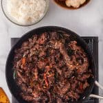 Overhead shot of a frypan cooking the last step of Ginger Beef. The fry pan is filled with crispy strips of beef in a dark brown sauce. To the top left of the fry pan is a bowl of white rice, and to the upper right of the frypan is a wooden bowl filled with onions, garlic, and ginger.