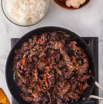 Overhead shot of a frypan cooking the last step of Ginger Beef. The fry pan is filled with crispy strips of beef in a dark brown sauce. To the top left of the fry pan is a bowl of white rice, and to the upper right of the frypan is a wooden bowl filled with onions, garlic, and ginger.