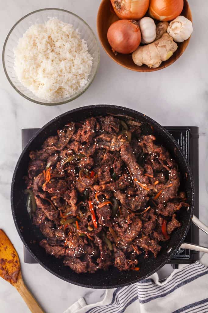 Overhead shot of a frypan cooking the last step of Ginger Beef. The fry pan is filled with crispy strips of beef in a dark brown sauce. To the top left of the fry pan is a bowl of white rice, and to the upper right of the frypan is a wooden bowl filled with onions, garlic, and ginger.