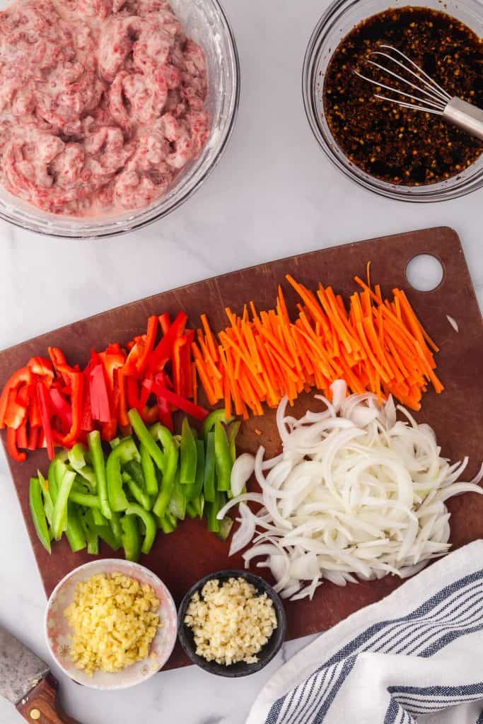 Overhead shot of veggies cut up on a cutting board which will be used in the ginger beef. Veggies include carrots, onions, green and red peppers, garlic and ginger. Top left corner is the bowl of the flank steak marinating in the cornstarch slurry. On the right is a small bowl of the sauce used in ginger beef.