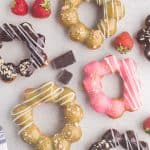 Overhead shot of 8 mochi donuts scattered on the counter. Four of them are coated in dark chocolate and sprinkled with slivered almonds, two have a pink glaze with some white ganache piped on one side and biscuit crumbs sprinkled on the other side. Two of them are dipped in a green tea ganache and have biscuits sprinkled on one side and white chocolate ganache piped on the other side.