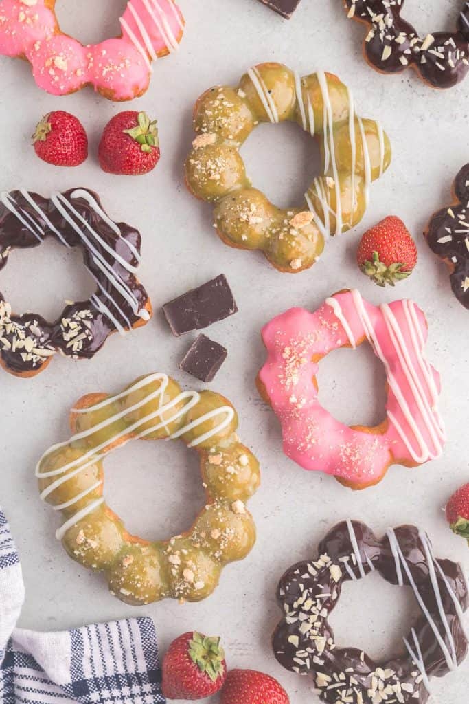 Overhead shot of 8 mochi donuts scattered on the counter.  Four of them are coated in dark chocolate and sprinkled with slivered almonds, two have a pink glaze with some white ganache piped on one side and biscuit crumbs sprinkled on the other side.  Two of them are dipped in a green tea ganache and have biscuits sprinkled on one side and white chocolate ganache piped on the other side.