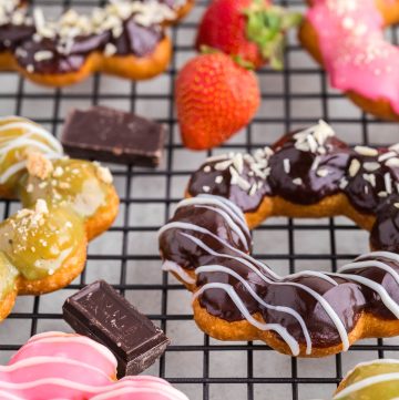 side angle shot looking up along a cooling rack filled with mochi donuts dipped in chocolate ganache, matcha ganache, and pink glaze. chunks of chocolate and fresh strawberries are lying as decoration between mochi donuts.