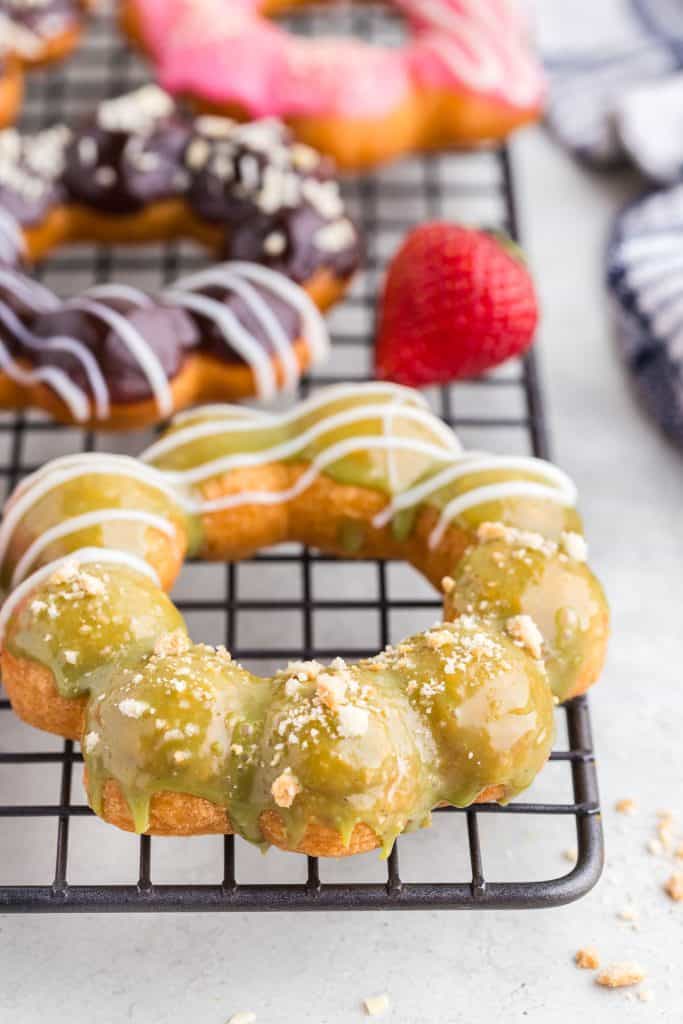 side view of a mochi donut that has been dipped in a matcha ganache, then one side sprinkled with cookie crumbs and the other side decorated with piped white chocolate.