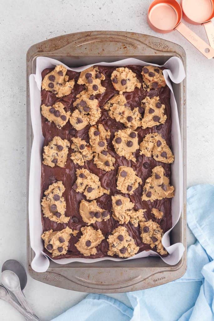 Overhead shot of the baking pan which was lined with parchment paper and then filled with the brownie batter. On top of the brownie batter are dollops of chocolate chip cookie dough scattered all around.