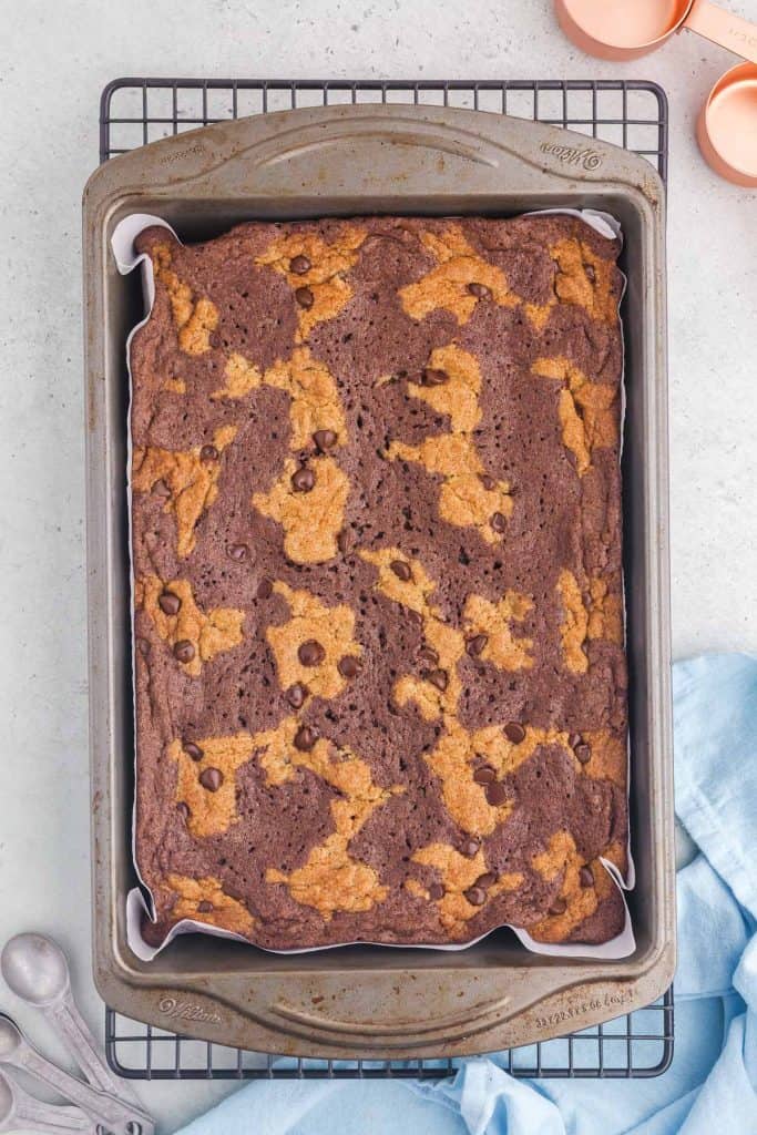 Overhead shot of the brookies after coming out of the oven. The baking pan is cooling on a cooling rack, there are a few copper measuring cups in the top right hand corner and a few aluminum measuring spoons in the bottom left hand corner, and a light blue tea towel in the bottom right corner.