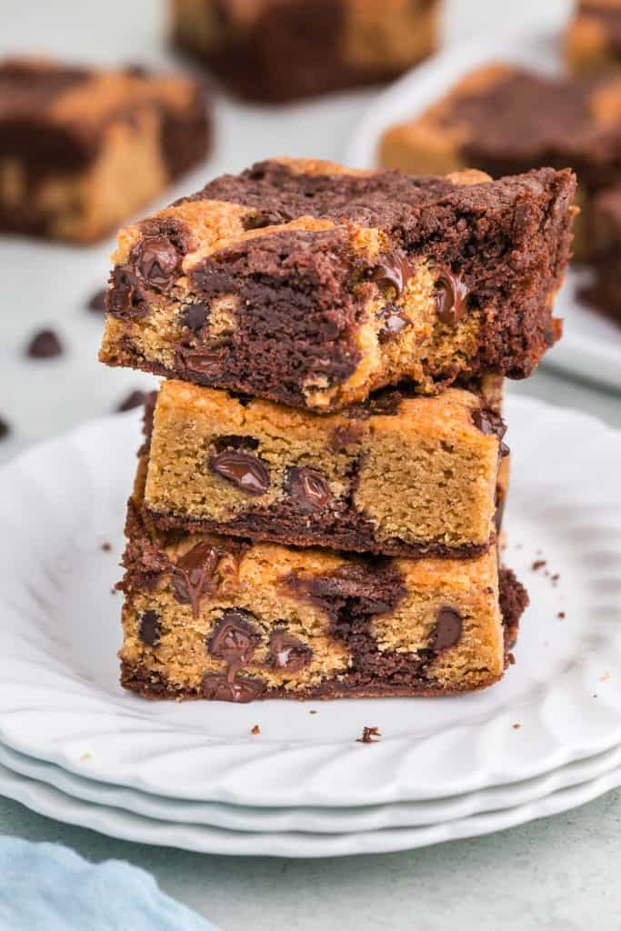 Side shot of three golden brown and marbled dark brown brookies stacked on top of each other on a white plate. The chocolate chips are melty warm and shiny. There are a few extra brookies in the background that are out of focus.