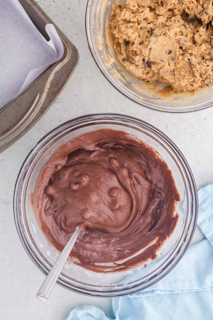 Overhead shot with the mixing bowl filled with the final brownie batter centered in the lower two thirds of the picture. In the upper right corner, the mixing bowl filled with cookie batter is peaking in to the scene, and the top left corner has the corner of the baking pan peeking in.