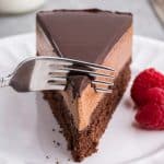 Close up side shot of a slice of chocolate mousse cake with it's three distinct layers of dark chocolate ganache, a lighter brown chocolate mousse and a darker brown chocolate cake layer. A fork is gently breaking in to the chocolate ganache layer to take a bite.