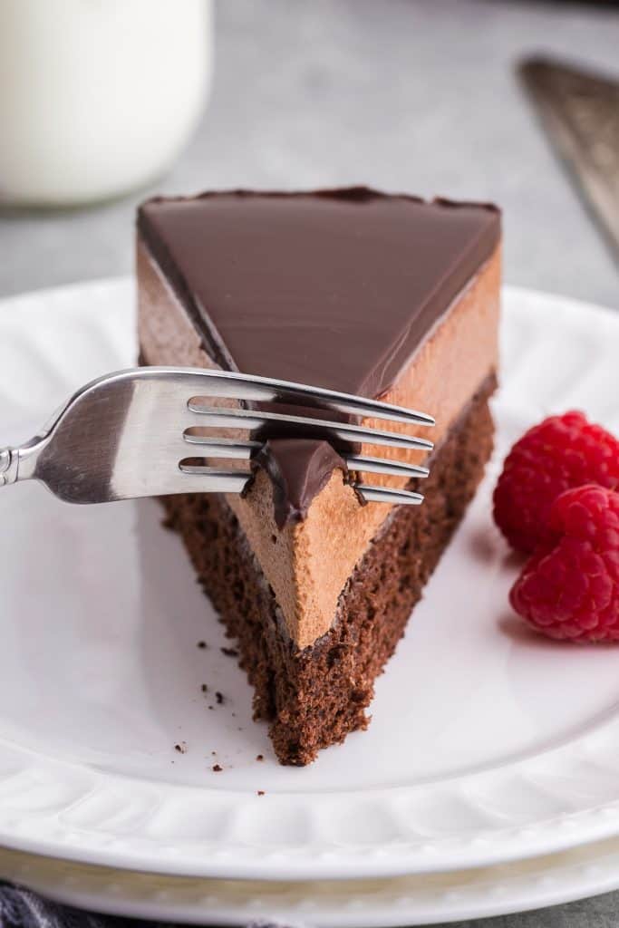 Close up side shot of a slice of chocolate mousse cake with it's three distinct layers of dark chocolate ganache, a lighter brown chocolate mousse and a darker brown chocolate cake layer. A fork is gently breaking in to the chocolate ganache layer to take a bite.