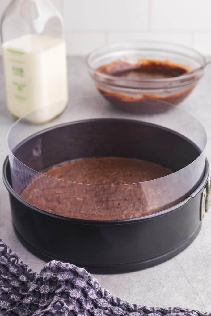 side view of the springform pan fitted with an acetate collar. Inside the pan is the chocolate cake, and around the edge and coming about an inch and a half over the top of the pan you can see the acetate film.