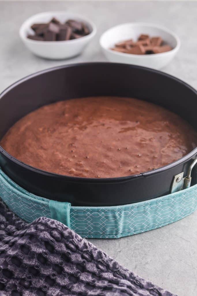Side shot of a springform pan filled with the chocolate chiffon cake batter and surrounded by a water soaked cake strip. In the back ground are two white bowls, one filled with dark chocolate and the other filled with milk chocolate.