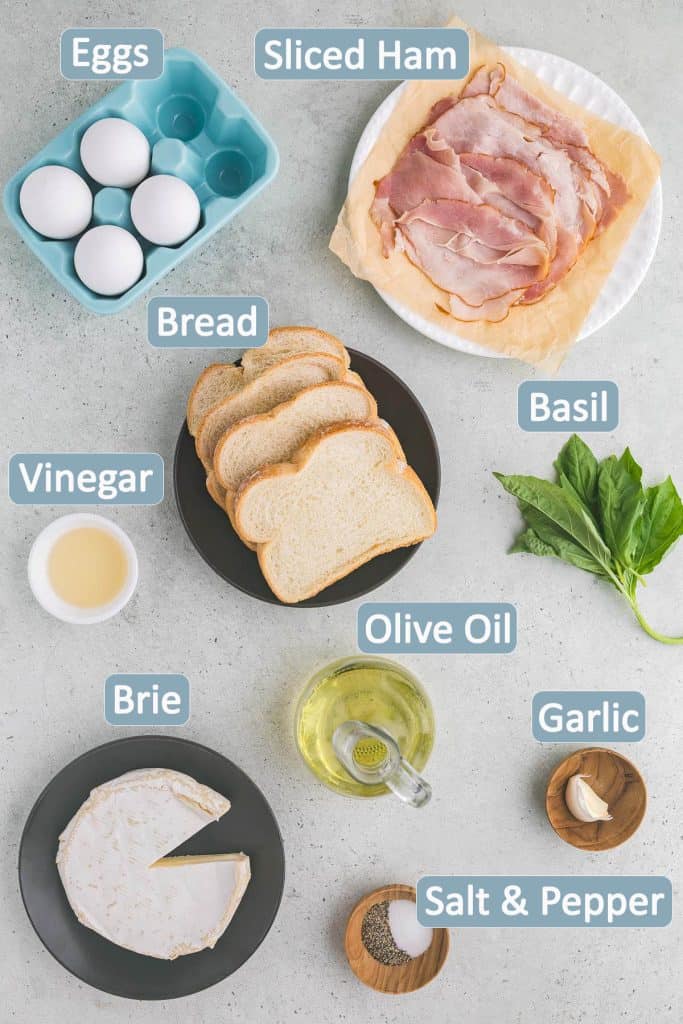 Overhead shot of the ingredients used to make this egg breakfast. Ingredients include: eggs, sliced ham, bread, vinegar, fresh basil, olive oil, brie cheese, garlic clove, salt and pepper.