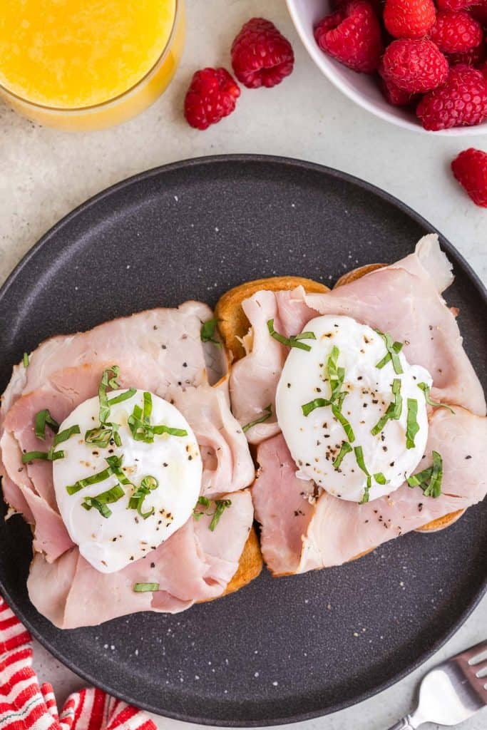 Overhead shot of a black plate with two slices of toast each rubbed with garlic, drizzled with olive oil, topped with brie cheese, ham and a poached egg, and garnished with fresh basil. The top left corner has a small glass of orange juice peeking in, and the top right hand corner has a small white bowl of fresh raspberries.