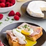 side view of a piece of toast topped with brie, ham, a poached egg, olive oil, salt, pepper, and fresh basil. The egg and toast have been cut in half releasing the runny yolk. In the back ground is a small white bowl of fresh raspberries, and a glass of orange juice.