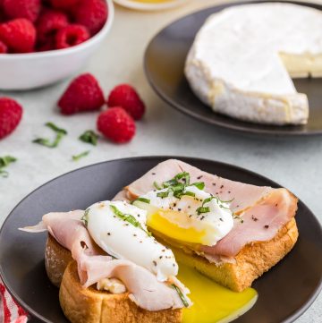 side view of a piece of toast topped with brie, ham, a poached egg, olive oil, salt, pepper, and fresh basil. The egg and toast have been cut in half releasing the runny yolk. In the back ground is a small white bowl of fresh raspberries, and a glass of orange juice.