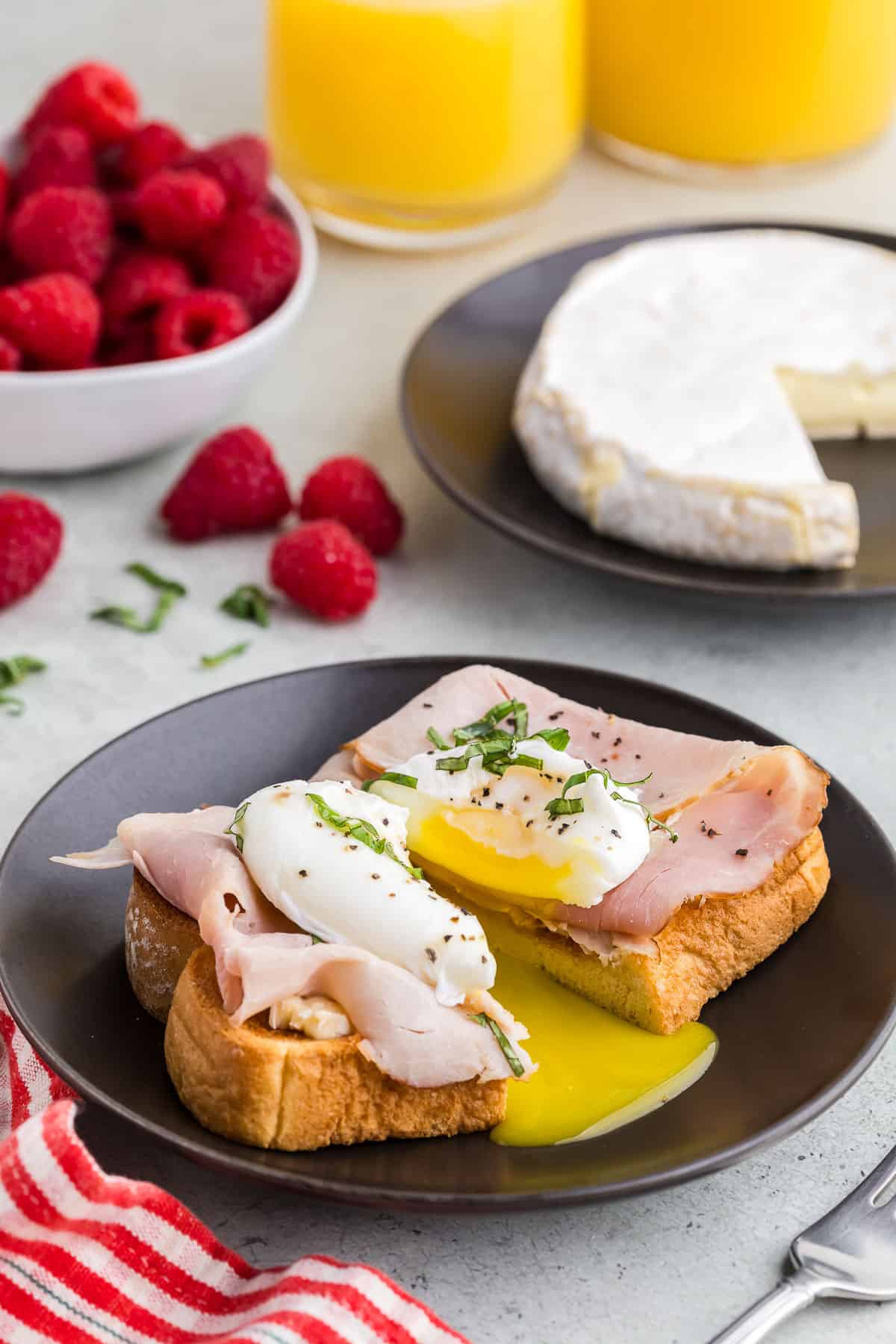 side view of a piece of toast topped with brie, ham, a poached egg, olive oil, salt, pepper, and fresh basil. The egg and toast have been cut in half releasing the runny yolk. In the back ground is a small white bowl of fresh raspberries, and a glass of orange juice.