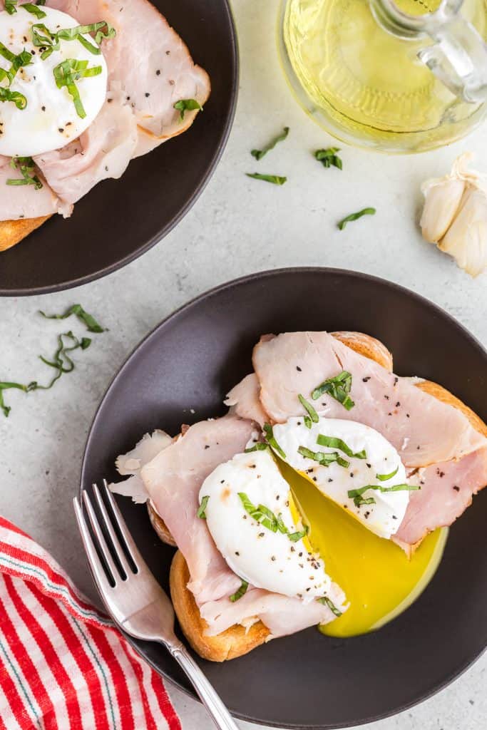 Overhead shot of a small black plate with a piece of toast covered in ham and a poached egg and garnished with basil. The egg, ham and toast has been cut in half and opened up to allow the runny yolk to spill out. The upper left corner has another plate of the egg breakfast peaking in. The right hand corner has a bulb of garlic and jar of olive oil peaking in.