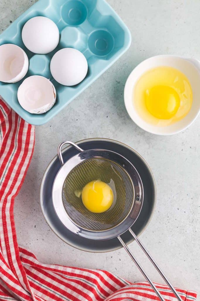 overhead shot of a fine mesh sieve sitting atop of small bowl. An egg has been cracked into the sieve to allow the watery part of the egg white to run into the bowl, leaving behind the good part of the egg white and yolk which will be poached. In the upper left corner of the photo is a small blue ceramic egg holder with 4 eggs in it. In the upper right corner of the photo is a small bowl with an egg cracked into it.