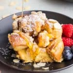 Side shot of a slice of baked French toast brioche on a small black plate garnished with raspberries and blueberries. A stream of syrup is coming down from the top and being poured on top of the French toast.