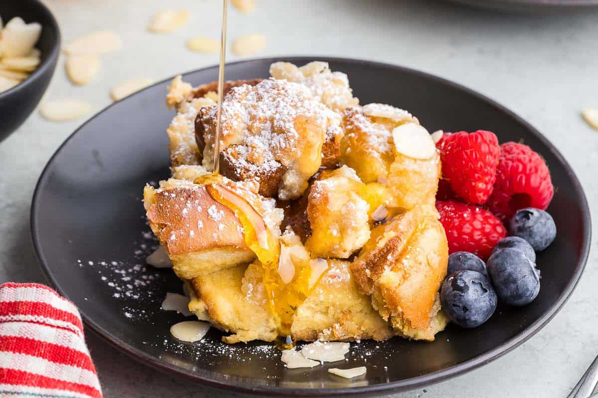 Side shot of a slice of baked French toast brioche on a small black plate garnished with raspberries and blueberries. A stream of syrup is coming down from the top and being poured on top of the French toast.
