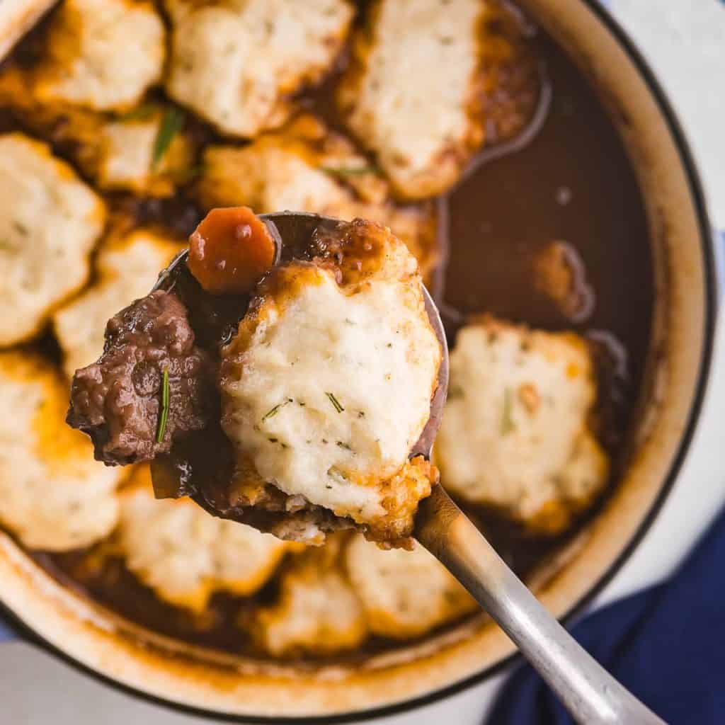 Overhead shot looking into a Dutch oven filled with beef stew and covered in rosemary dumplings. A ladle is bringing a large scoop up towards the camera with a large dumpling and a juicy chunk of beef next to it.