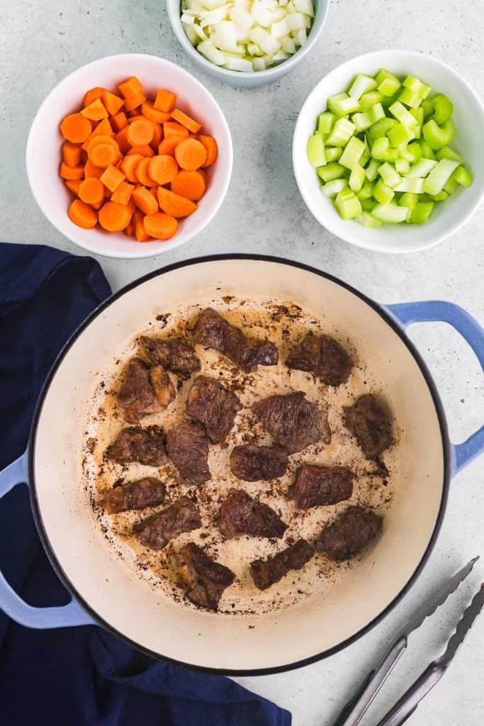 Overhead shot of a Dutch oven with beef cubes searing. Above the Dutch oven in the photo there are bowls of carrots, onion, and celery ready to be added next.