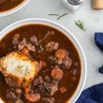 Overhead shot of a white bowl in the lower left corner filled with beef stew and a rosemary dumpling. The stew is a rich brown and filled with large chunks of beef, carrots, and celery. There is another bowl of stew peeking in in the upper left corner, and on the right are salt and pepper shakers, along with a wine cork peeking in. there is some rosemary scattered around the upper right corner.