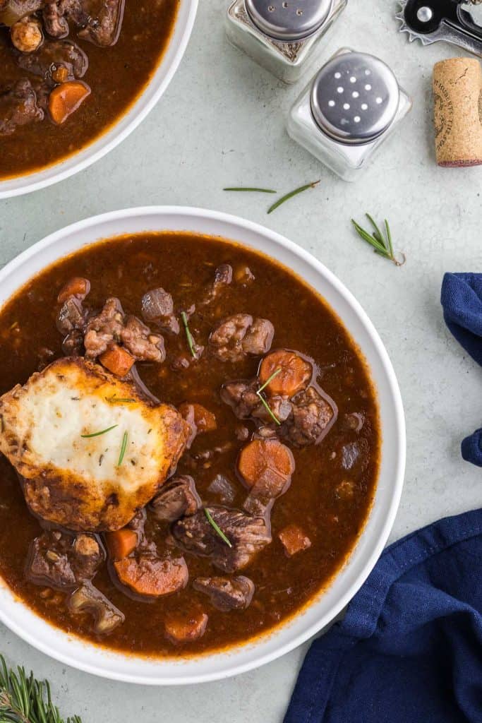 Overhead shot of a white bowl in the lower left corner filled with beef stew and a rosemary dumpling. The stew is a rich brown and filled with large chunks of beef, carrots, and celery. There is another bowl of stew peeking in in the upper left corner, and on the right are salt and pepper shakers, along with a wine cork peeking in. there is some rosemary scattered around the upper right corner.